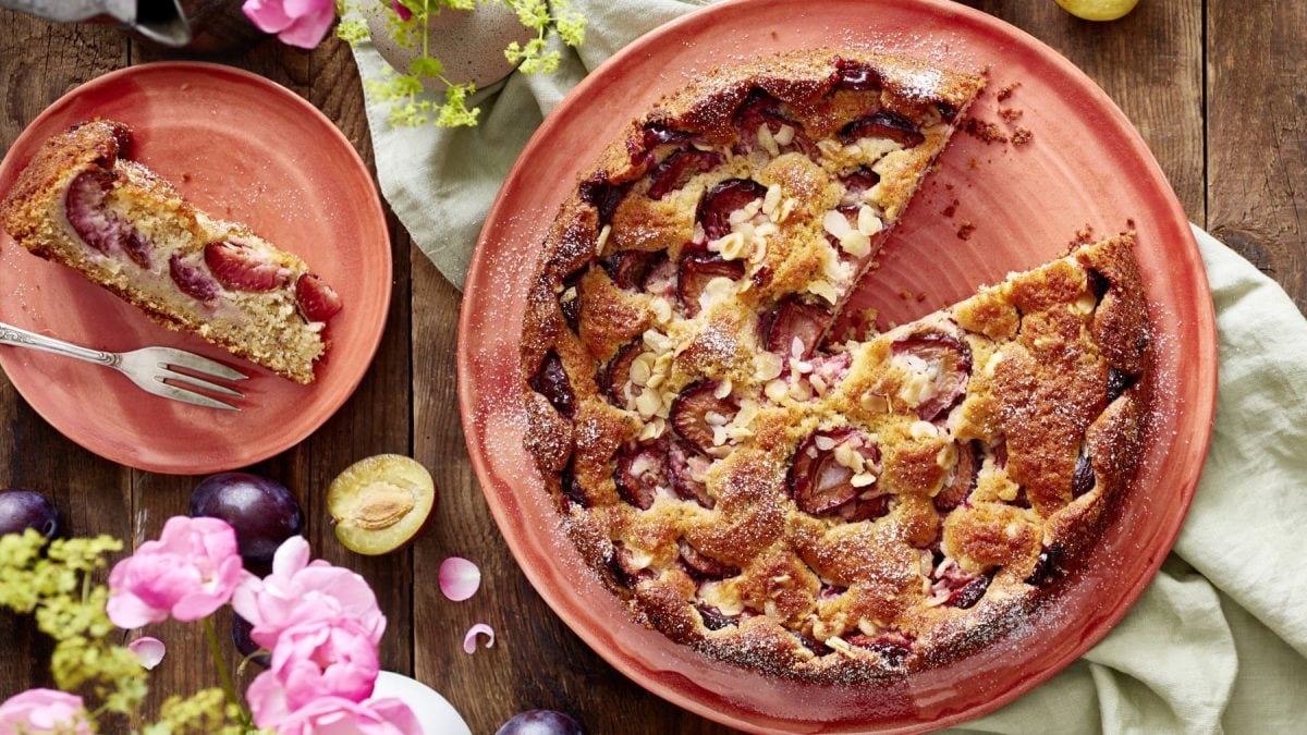 Ein versunkener Pflaumenkuchen auf einem hellroten Servierteller auf einem grünen Geschirrtuch auf einem hölzernen Hintergrund. Links davon ein Teller mit einem Stück Kuchen und links unten Blumen und Pflaumen.