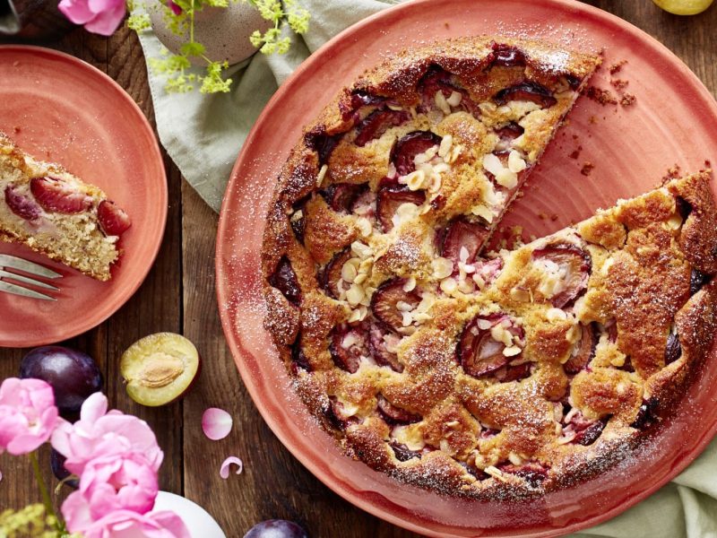 Ein versunkener Pflaumenkuchen auf einem hellroten Servierteller auf einem grünen Geschirrtuch auf einem hölzernen Hintergrund. Links davon ein Teller mit einem Stück Kuchen und links unten Blumen und Pflaumen.