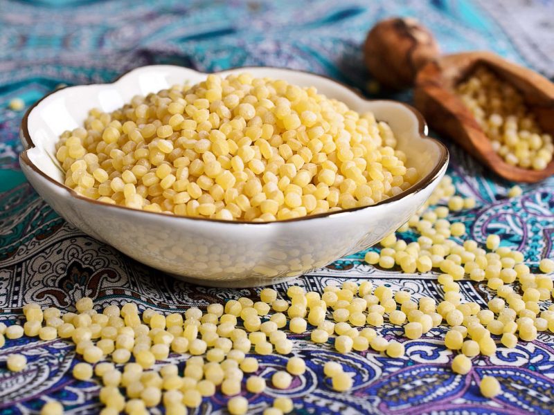 Auf einem orientalisch angehauchtem Untergrund steht eine Schüssel Ptitim. Der israelische Couscous ist zudem drum herum verstreut.