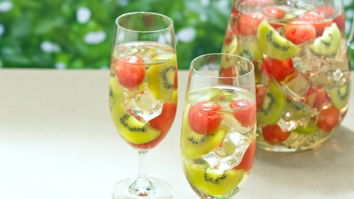 White Sangria mit Melone und Kiwi in einer Schale und zwei gefüllte Sektgläser auh hellem Tisch. Außenaufnahme. Frontalansicht.