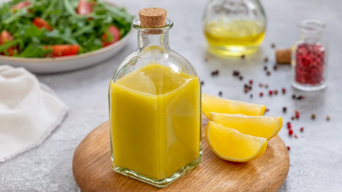 Eine Glasflasche mit dem Zitronendressing auf einem hellen Tisch mit rotem Pfeffer und einem Teller mit Salat.