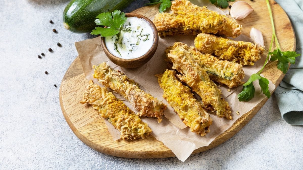 Ein Holzbrett mit den Zucchini-Sticks auf grauem Untergrund mit Zucchini, Knoblauch und frischer Petersilie.