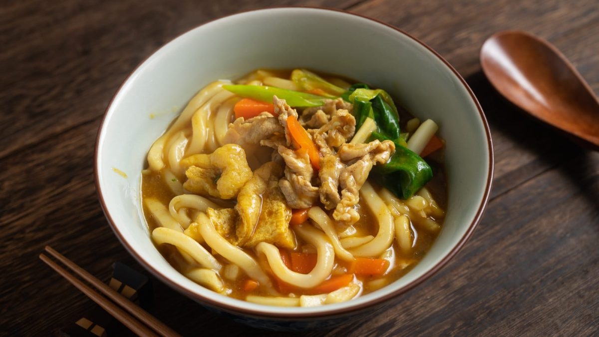Eine Schüssel mit Curry Udon neben Holzbesteck auf einem Holztisch.