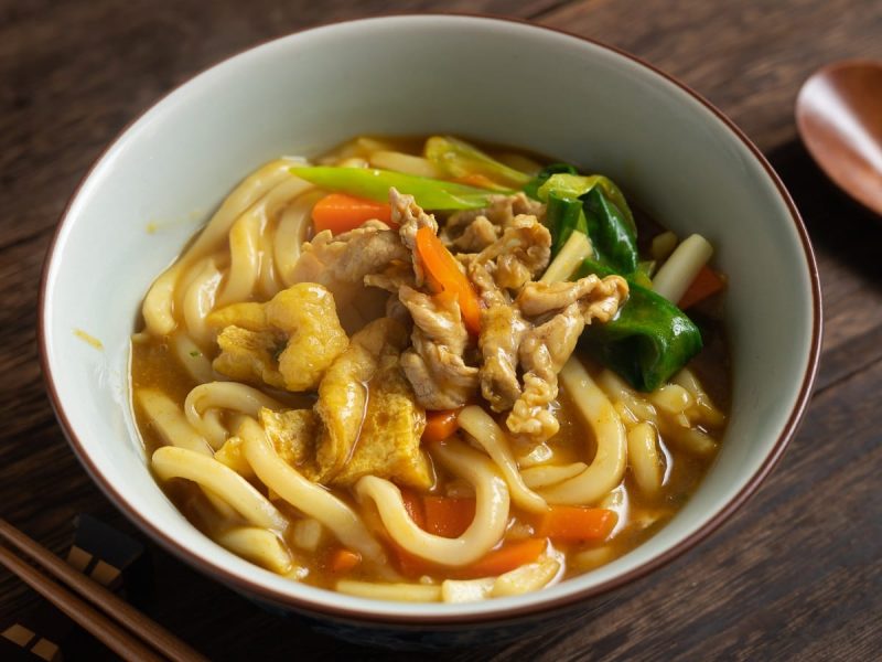 Eine Schüssel mit Curry Udon neben Holzbesteck auf einem Holztisch.