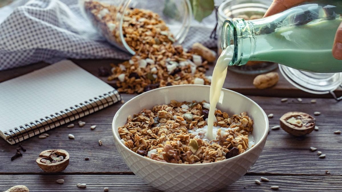 Frontalsicht: Eine Schüssel mit Apfel-Nuss-Granola steht auf einem Tisch. Eine weibliche Hand hält eine Glasflasche Milch in der Hand und schüttet Milch in die Schahle. Drum herum liegen ein Küchenhandtuch und ein Schreibblock.