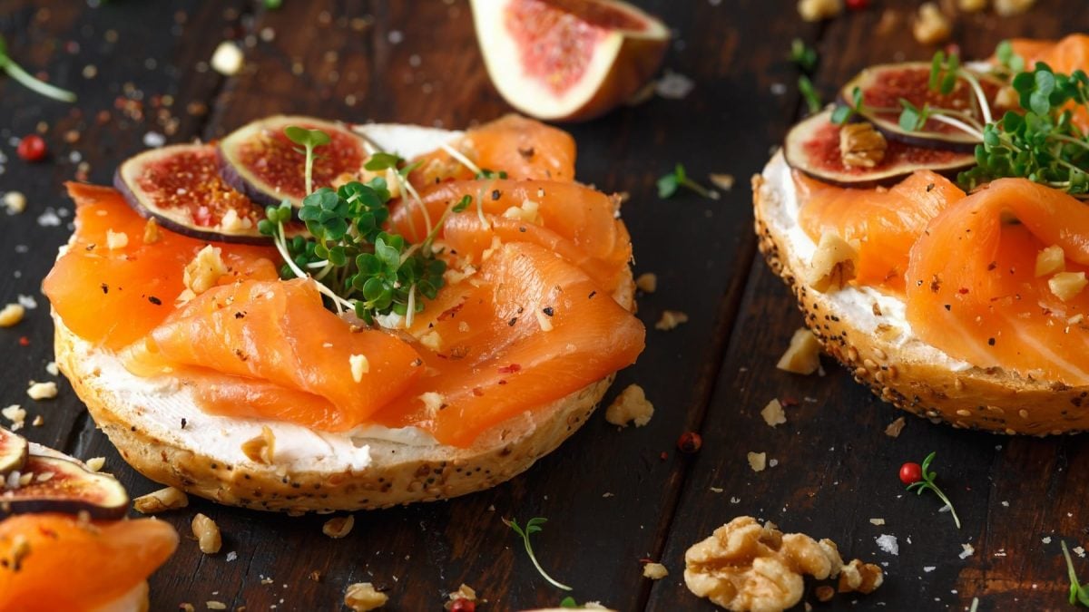 Zwei Bagelhälften belegt mit Lachs, Feigen, Walnuss und Kresse auf einem dunkelbraunen Holztisch seitlich von schräg oben fotografiert.