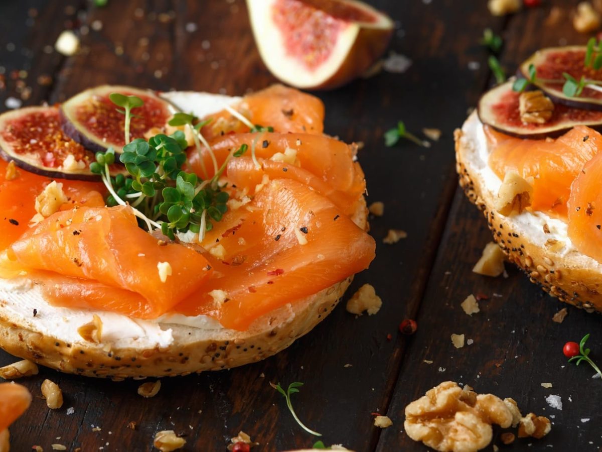 Zwei Bagelhälften belegt mit Lachs, Feigen, Walnuss und Kresse auf einem dunkelbraunen Holztisch seitlich von schräg oben fotografiert.