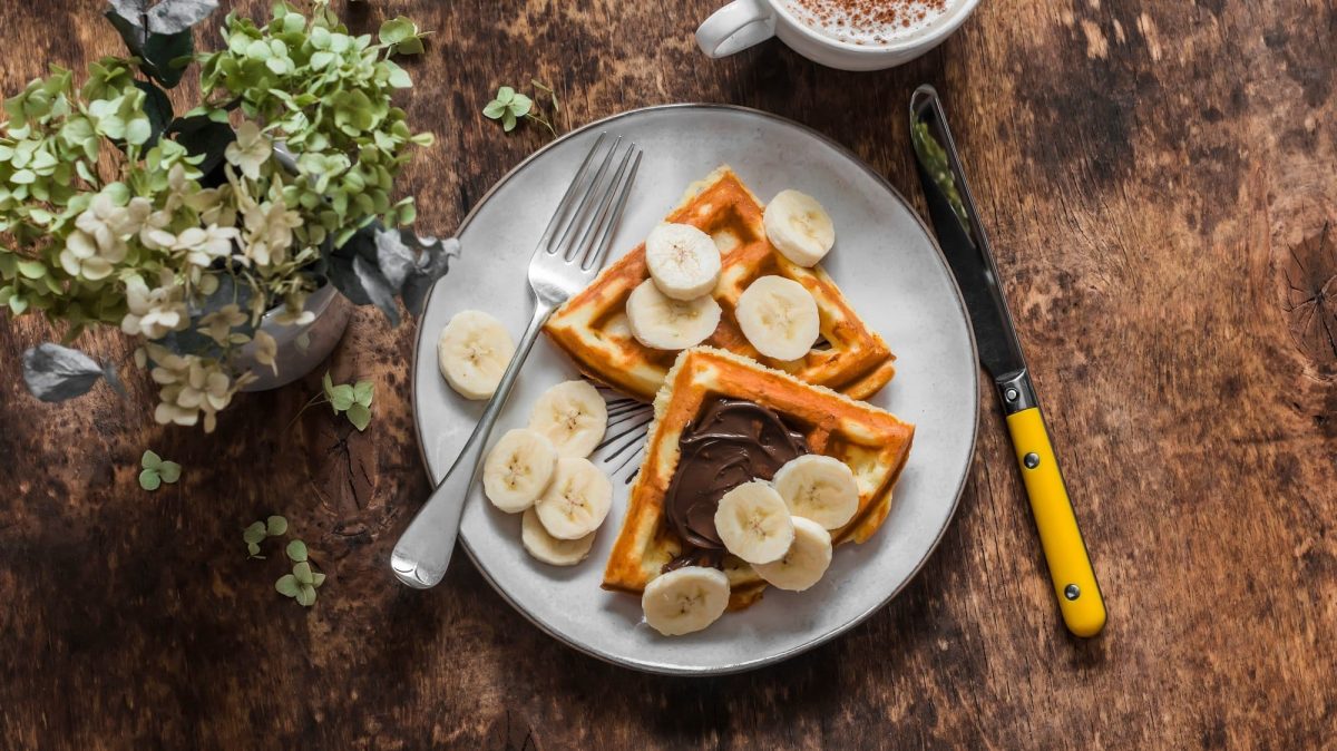 Ein Teller mit 2 Bananenbrot-Waffeln, frischen Bananenscheiben und Schokocreme in der Draufsicht, daneben Besteck und eine Tassee Cappuccino sowie Blumen.