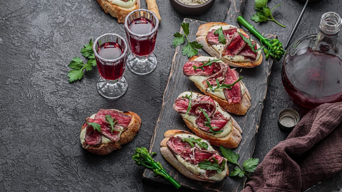 Draufsicht: Dunkler Untergrund und ein Holzbrettchen mit mehreren Beef-Bruschette mit Avocado-Creme. Daneben stehen eine Karaffe und eine Glaskaraffe mit Rotwein.