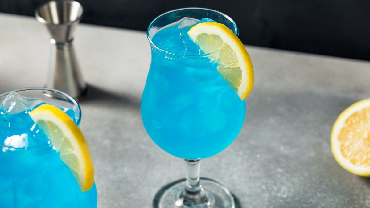 Ein Glas mit dem blauen Cocktail auf einem grauen Tisch mit schwarzem Hintergrund. Alles seitlich fotografiert.
