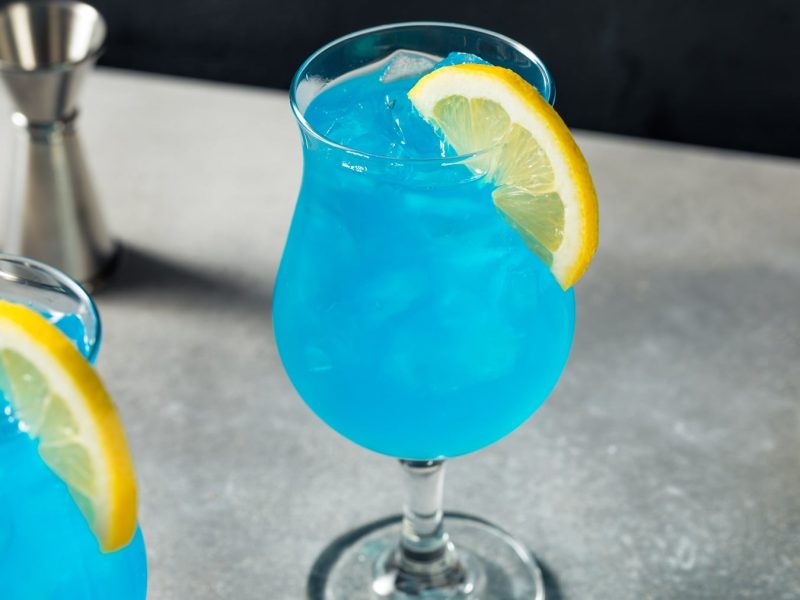 Ein Glas mit dem blauen Cocktail auf einem grauen Tisch mit schwarzem Hintergrund. Alles seitlich fotografiert.