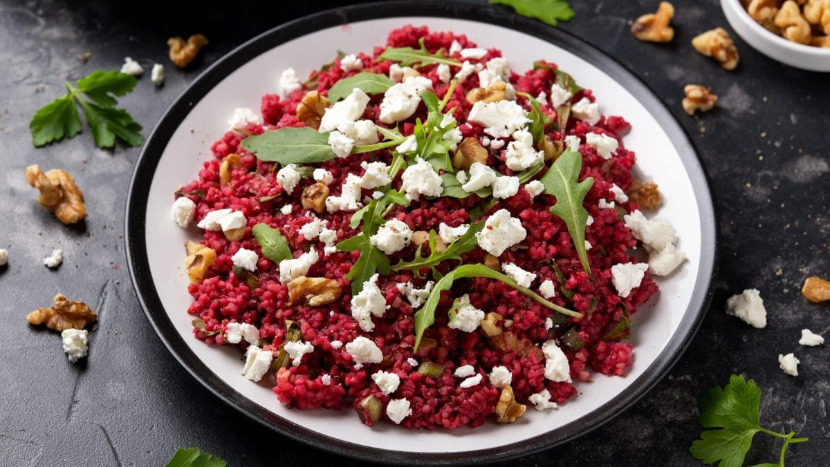 Draufsicht: ein Teller mit Bulgur mit Rote Bete und Walnüssen auf einem dunklen Holztisch. Drum herum sind Walnüsse und Ruccola-Blätter verstreut.