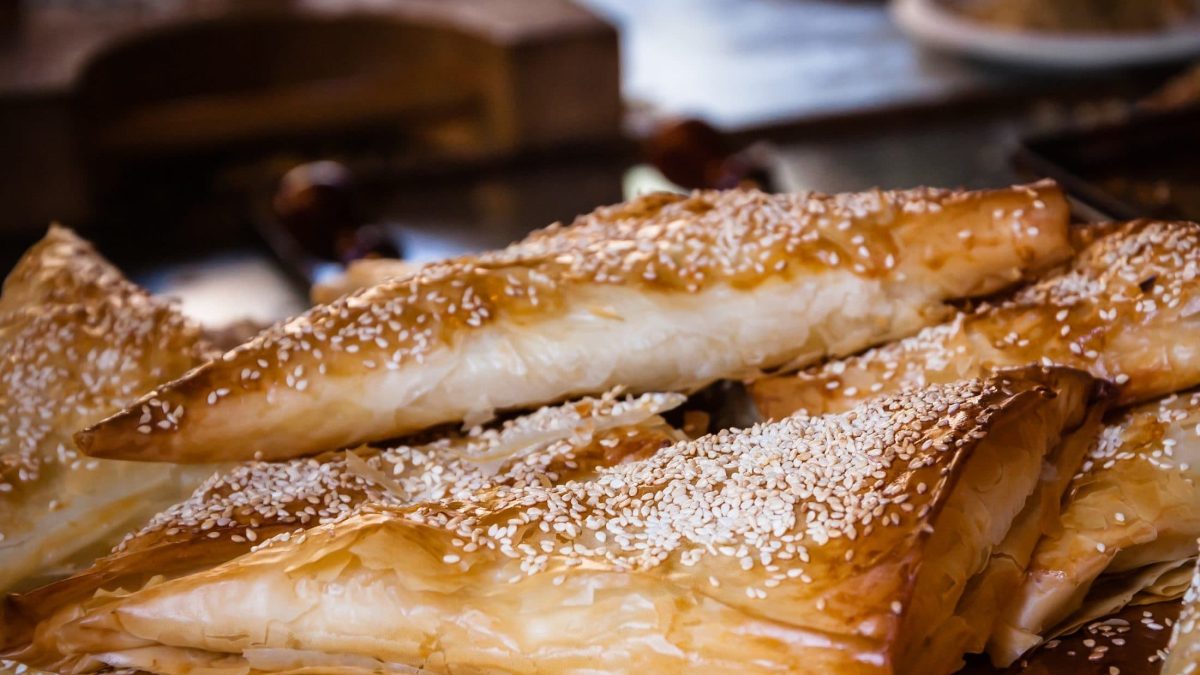 Frontal: dreieckige, im Ofen gebackene Teigtaschen, liegen aufeinander. Es handelt sich um Bureka, die mit weißem Sesam bestreut sind.