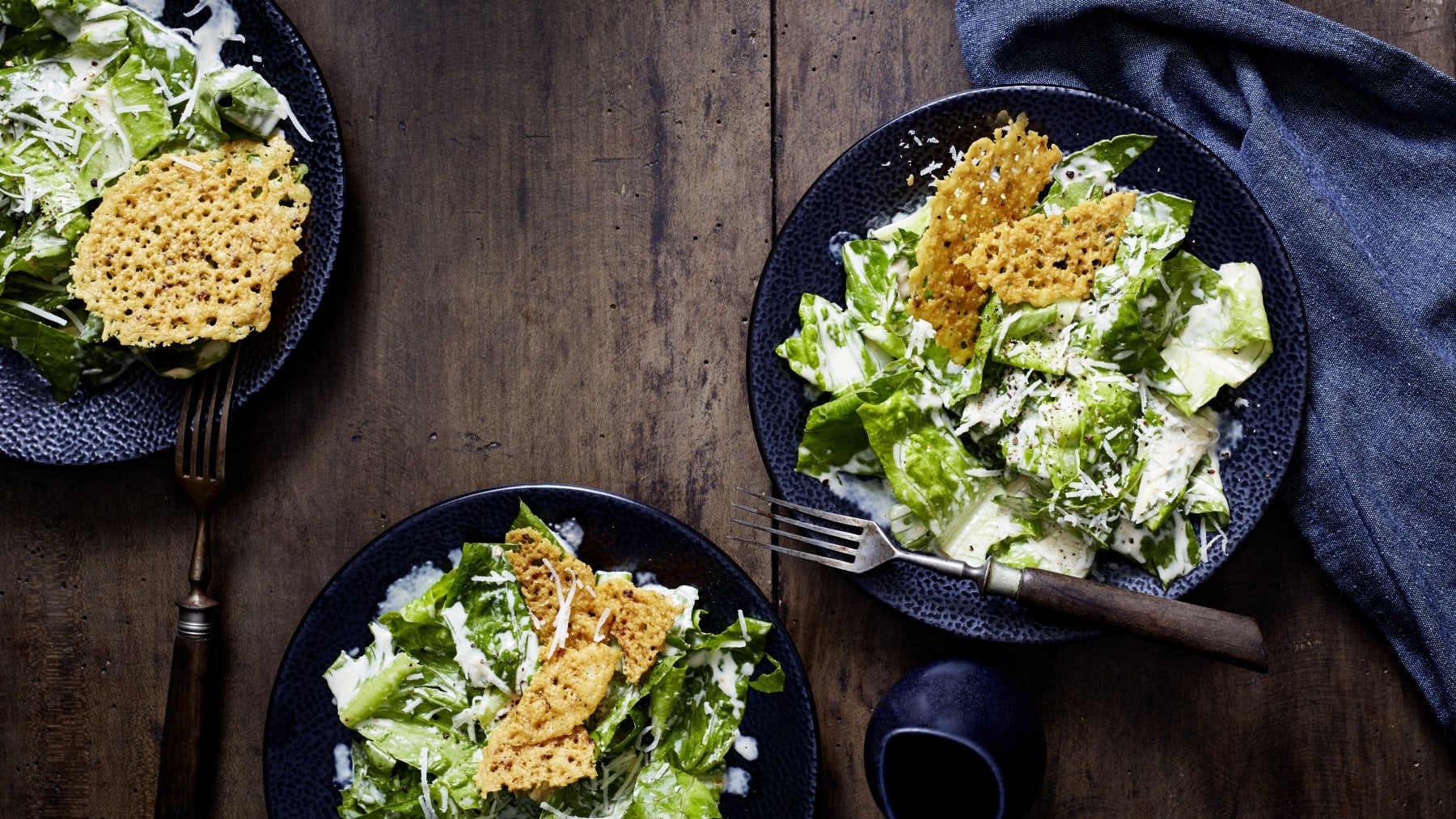 Drei schwarze Teller auf einem hölzernen Hintergrund mit Caesar-Greens und Parmesanbruch und Gabeln. Rechts davon ein schwarzes Geschirrtuch.