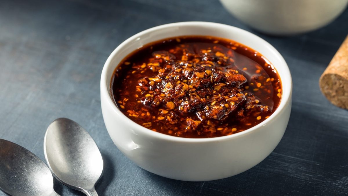 Eine weiße Schüssel mit Garlic-Chili-Crisp auf einem grauen Hintergrund. Links unten zwei Löffel.