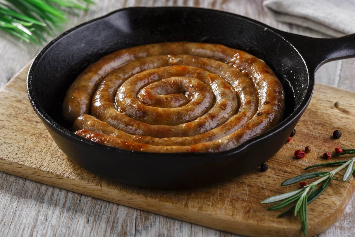 Draufsicht, schräg: eine schwarze gusseiserne Pfanne in der eine gebratene, schneckenförmige, runde Bratwurst liegt. Es handelt sich um eine Englische Wurst-Spezialität. Das ist eine Englische Wurst-Spezialität.