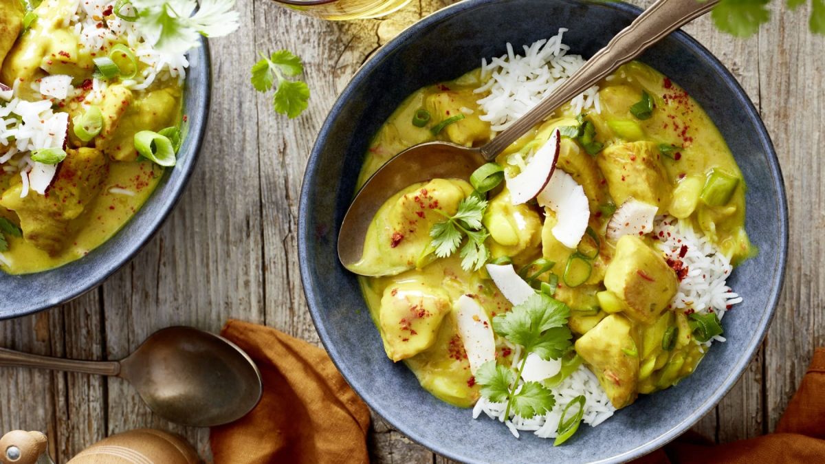 Zwei Portionen Curry-Putengulasch in grauen Schüsseln mit Löffeln auf einem hölzernen Hintergrund. Unten noch ein Löffel, ein oranges Geschirrtuch und eine Pfeffermühle.
