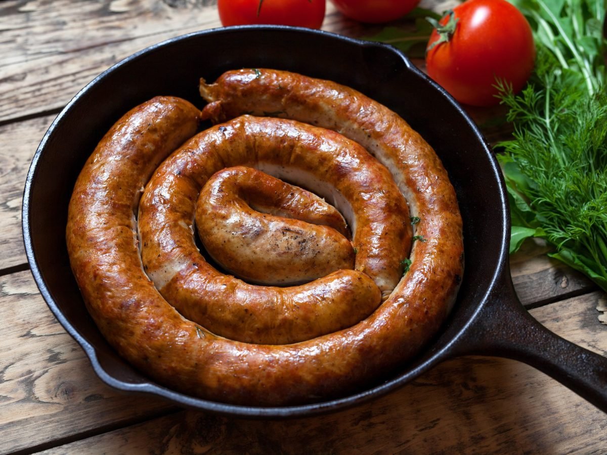 Draufsicht, schräg: eine schwarze gusseiserne Pfanne in der eine gebratene, schneckenförmige, runde Bratwurst liegt. Es handelt sich um eine Englische Wurst-Spezialität. Das ist eine Englische Wurst-Spezialität.