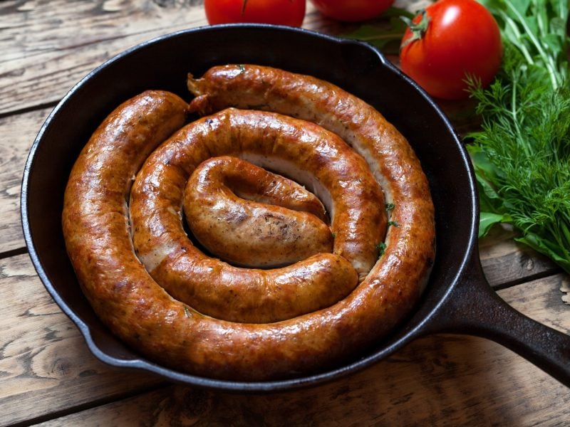 Draufsicht, schräg: eine schwarze gusseiserne Pfanne in der eine gebratene, schneckenförmige, runde Bratwurst liegt. Es handelt sich um eine Englische Wurst-Spezialität. Das ist eine Englische Wurst-Spezialität.