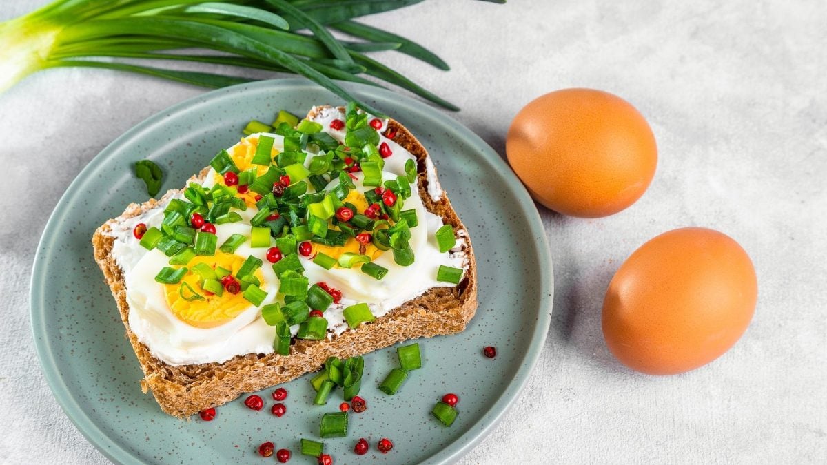 Draufsicht, schräg: ein blauer Teller mit einem Eier-Sandwich mit scharfem Hüttenkäse auf einem hellen Untergrund. Daneben liegen zwei intakte Eier und etwas Schnittlauch.