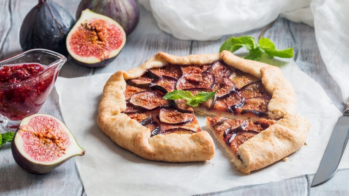 Eine angeschnittene einfache Feigen-Galette auf einem Stück Backpapier, daneben ein Messer, frische, zum Teil aufgeschnittene Feigen und ein Krug mit Feigenmarmelade.