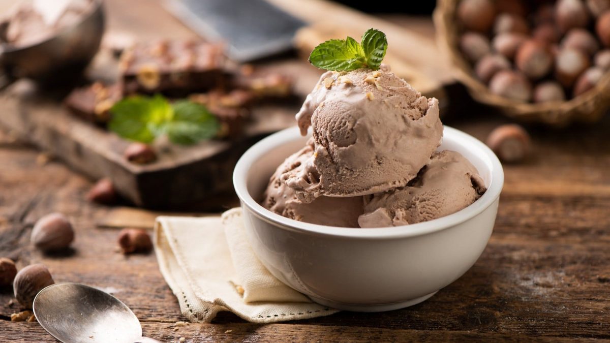 eine Schüssel mit 3 Kugeln Nutella-Eis, garniert mit Minze, daneben Haselnüsse und ein Löffel.