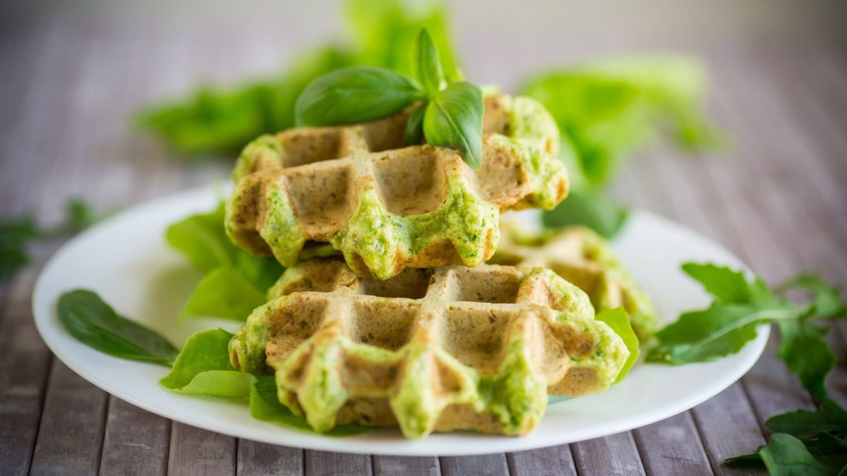 Frontalsicht. Zwei Falafel-Waffeln auf einem weißen Teller. Drum herum liegen frische Kräuter.