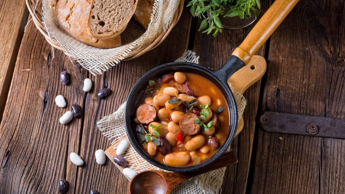 Draufsicht: rustikaler Holztisch mit einer Pfanne Fasolka po bretonsku. Es handelt sich um eine polnischen Bohnen-Würstchenpfanne. Daneben liegt ein angeschnittenes Brot.