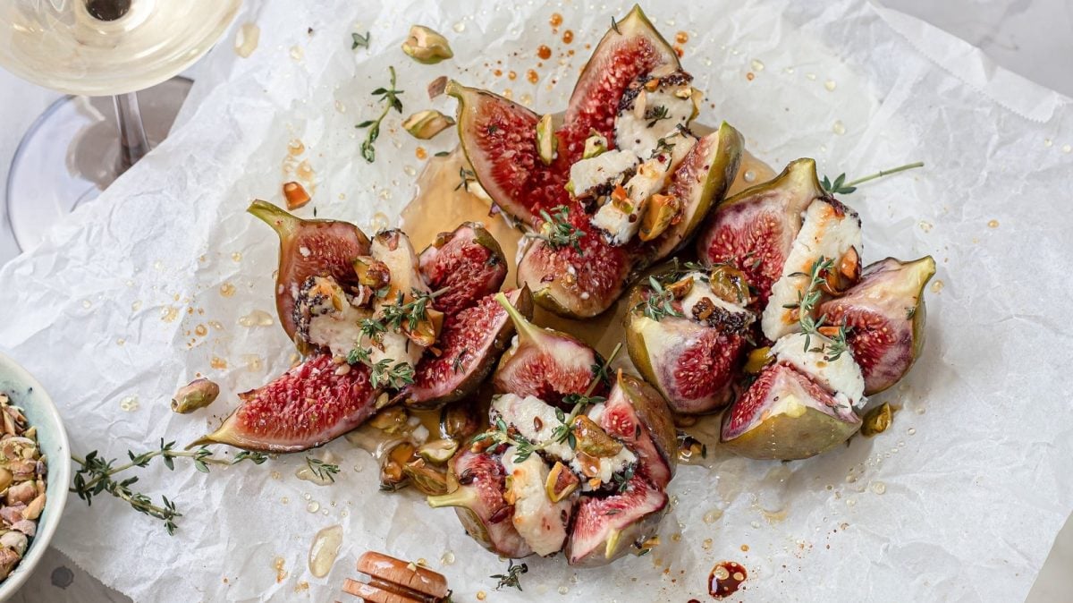 Gebackene Feigen mit Ziegenkäse auf weißem Backpapier. Links unten eine kleine Schüssel mit Pistazien und links oben ein Glas Weißwein.