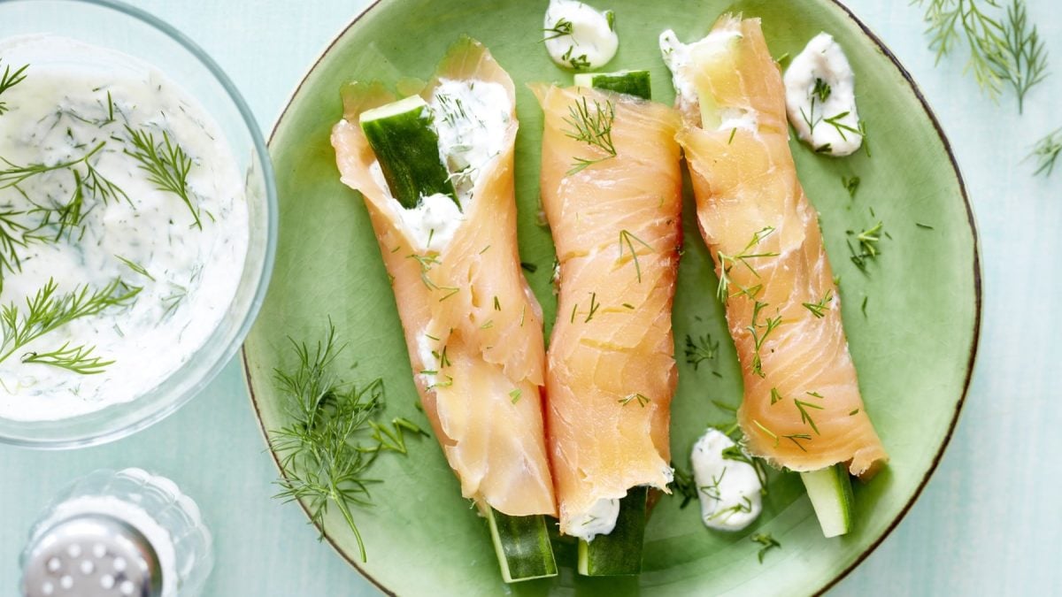 Drei Gefüllte Lachsröllchen auf grünem Teller, daneben weißer Dip mit Dill und Salzstreuer auf hellem Untergrund.