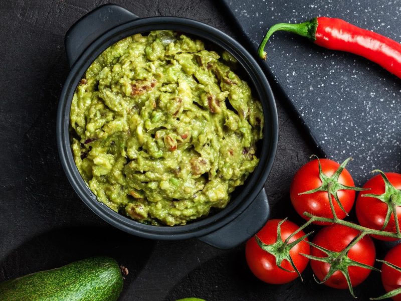 Eine schwarze Schüssel mit der Guacamole auf dunklem Untergrund mit Chili, Tomaten und Limette.