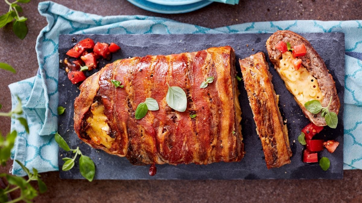 Angeschnittener Hackbraten mit Makkaroni-Füllung auf schwarzem Brett, darunter blaues Küchentuch, dunkler Untergrund und Kräuter am Rand.