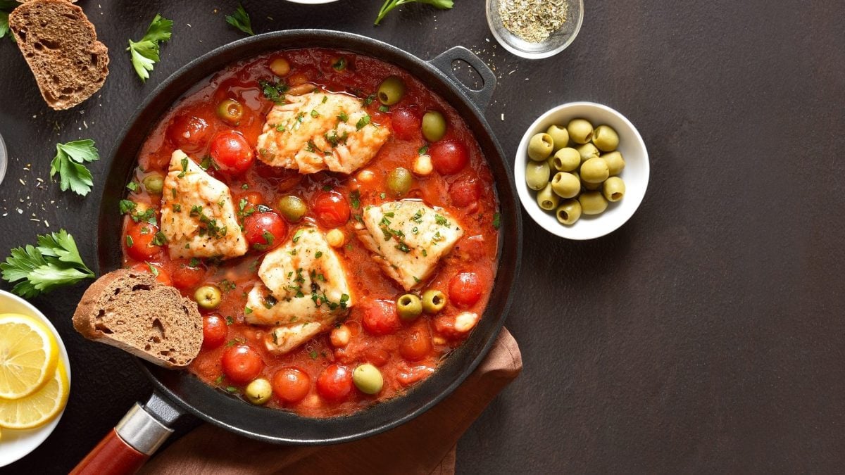 Draufsicht: Eine Kabeljau-Pfanne mit Tomaten, Oliven und Kichererbsen steht auf einem dunklen Untergrund. Drum herum sind Schälchen mit frischen Zutaten und Kräuter.