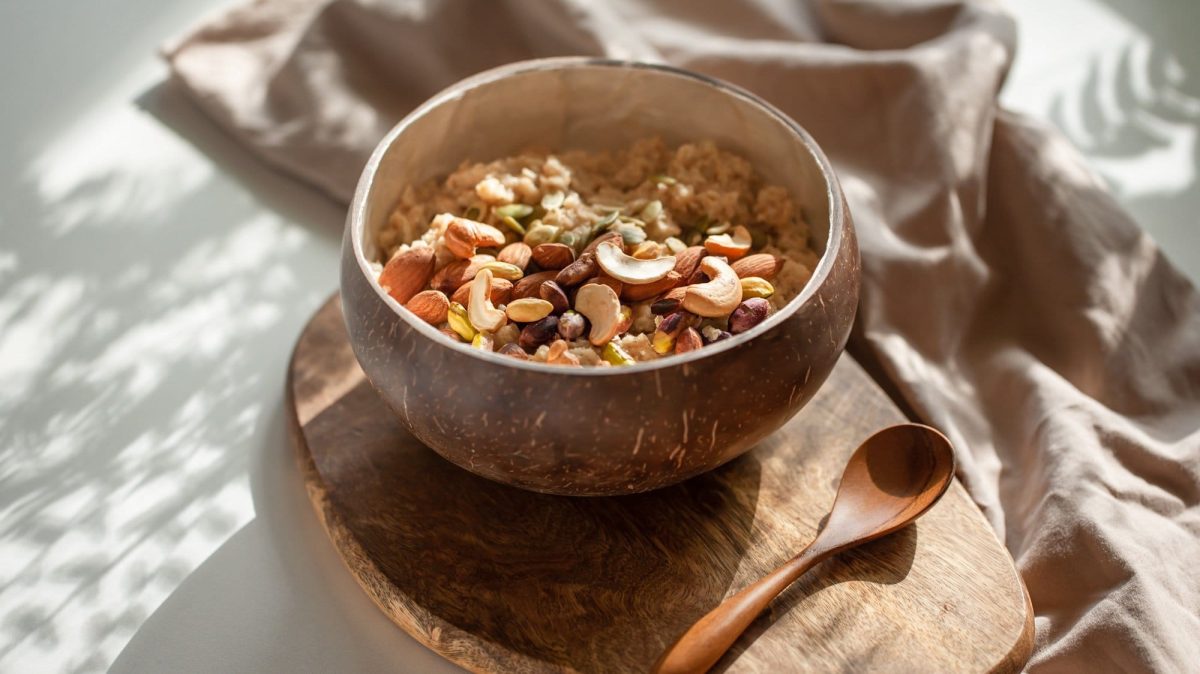 Eine Holzschale Kamillen-Porridge mit Nüssen garniert auf einem Holzbrett, daneben ein Holzlöffel, Schatten von Sonnenstrahlen.