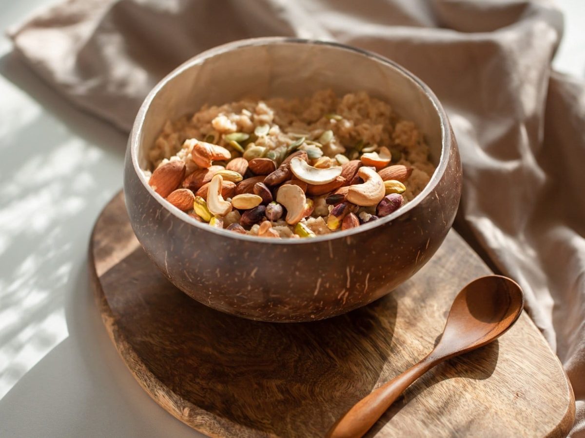 Eine Holzschale Kamillen-Porridge mit Nüssen garniert auf einem Holzbrett, daneben ein Holzlöffel, Schatten von Sonnenstrahlen.