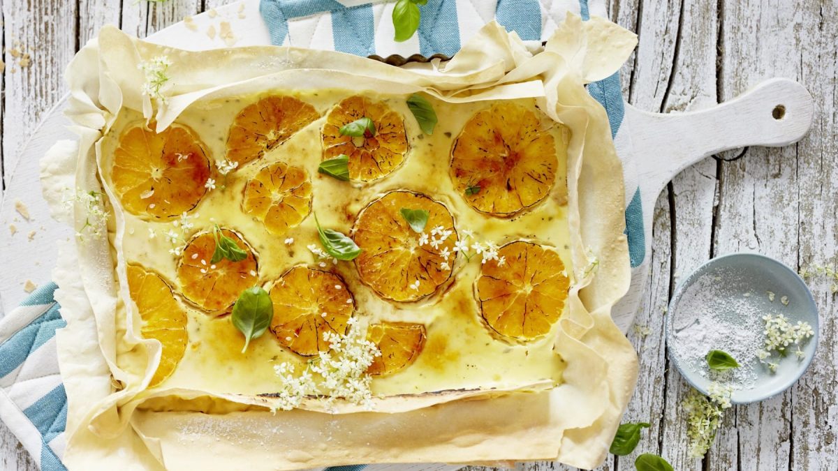 Eine karamellisierte Orangen-Tarte auf blau-weißen Ofentüchern auf einem hölzernen Hintergrund. Rechts davon ein Schälchen mit Blüten und Puderzucker.