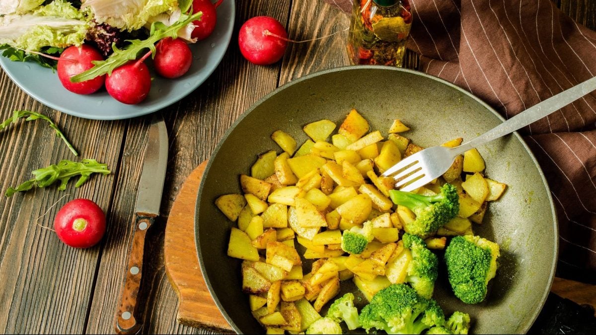 Draufsicht: Untergrund aus Holz, eine Kartoffel-Brokkoli-Pfanne und drum herum liegen Radieschen. Daneben ist eine Schüssel Salat.