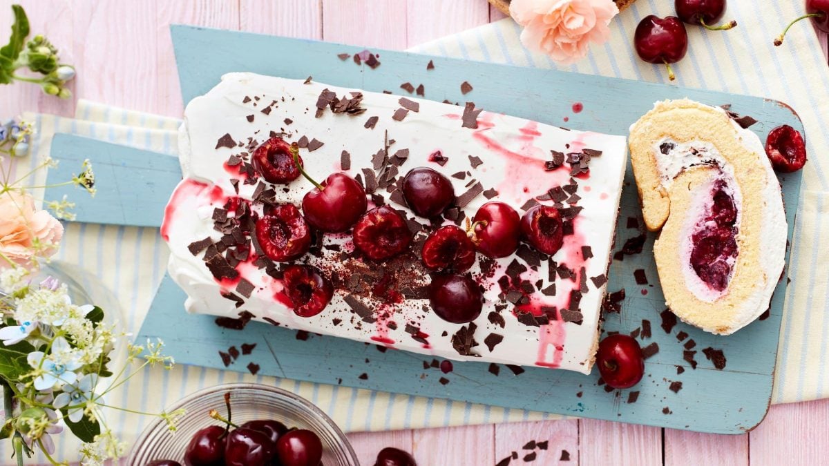 Ein Kirsch-Käsekuchen-Biskuit auf einem blauen Brett auf einem blau, gelb gestreiften Geschirrtuch. Der Hintergrund ist rosa. Links von dem Biskuit Blumen und unten eine kleine Schüssel mit Kirschen. Rechts oben Kirschen und eine Rose.