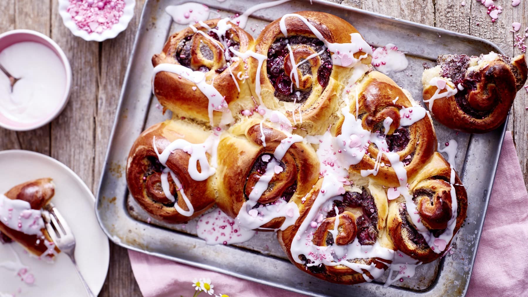 Kirsch-Mohn-Schnecken auf einem silbernen Blech auf einem hölzernen Hintergrund. Links davon ein kleiner Teller mit einer Kirsch-Mohn-Schnecke, eine kleine Schüssel mit Zuckerguss und Zuckerstreusel. Rechts ein rosarotes Geschirrtuch.