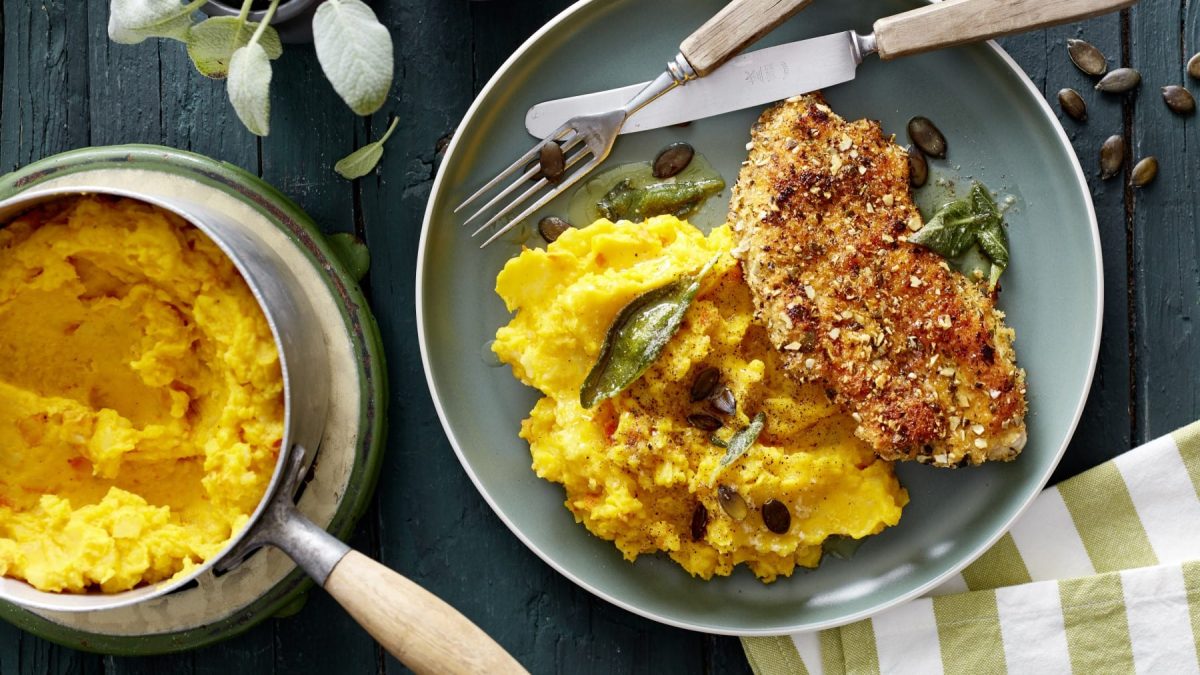 Ein Teller mit Knusper-Schnitzel mit Kürbisstampf und Besteck, daneben Topf mit Kürbisbrei auf dunklem Untergrund.