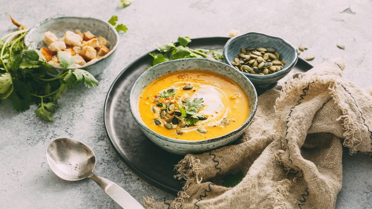 Eine Schüssel Kürbis-Curry-Suppe mit Kürbiskernen, Kräutern und Schmand, daneben ein Löffel, ein Geschirrtuch, eine Schale mit Kürbiskernen und Croûtons.