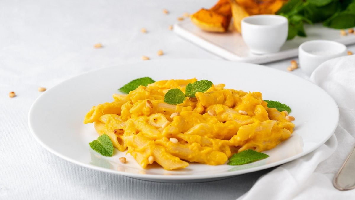 Ein Teller Kürbis-Feta-Pasta mit Minze und Pinienkernen garniert, dahinter unscharf gebackene Kürbisspalten auf einem Teller.