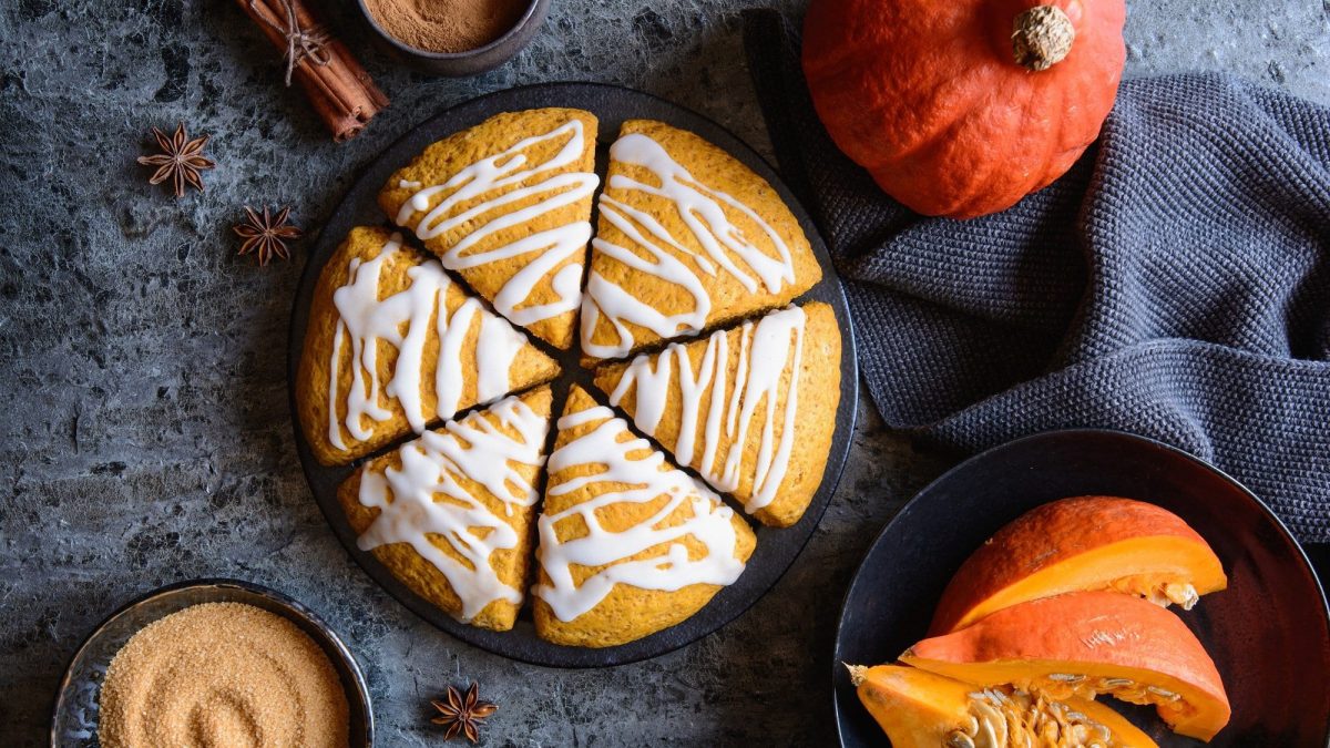 6 Kürbis-Scones wie eine Torte arrangiert auf einem Teller in der Draufsicht, drumherum ein ganzer und ein geschnittener Kürbis, eine Schale brauner Zucker, Zimstangen.