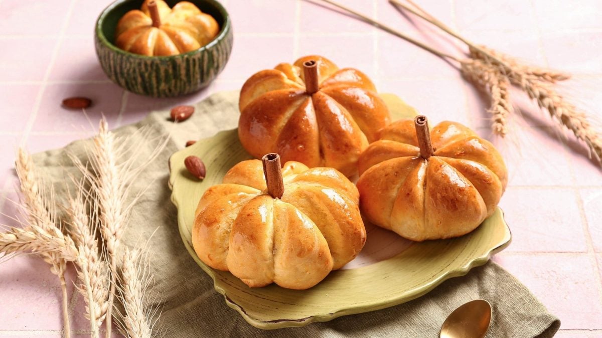 3 Kürbisbrötchen auf einem Teller, dahinter eine Schüssel mit einem weiteren Brötchen, außerdem drumherum Getreideähren.