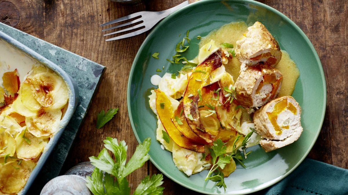 Eine Portion Kürbisröllchen mit Gratin auf einem türkisen Teller auf einem hölzernen Hintergrund. Links davon eine Auflaufform. Oben eine Gabel und unten eine Pfeffermühle und Petersilie.