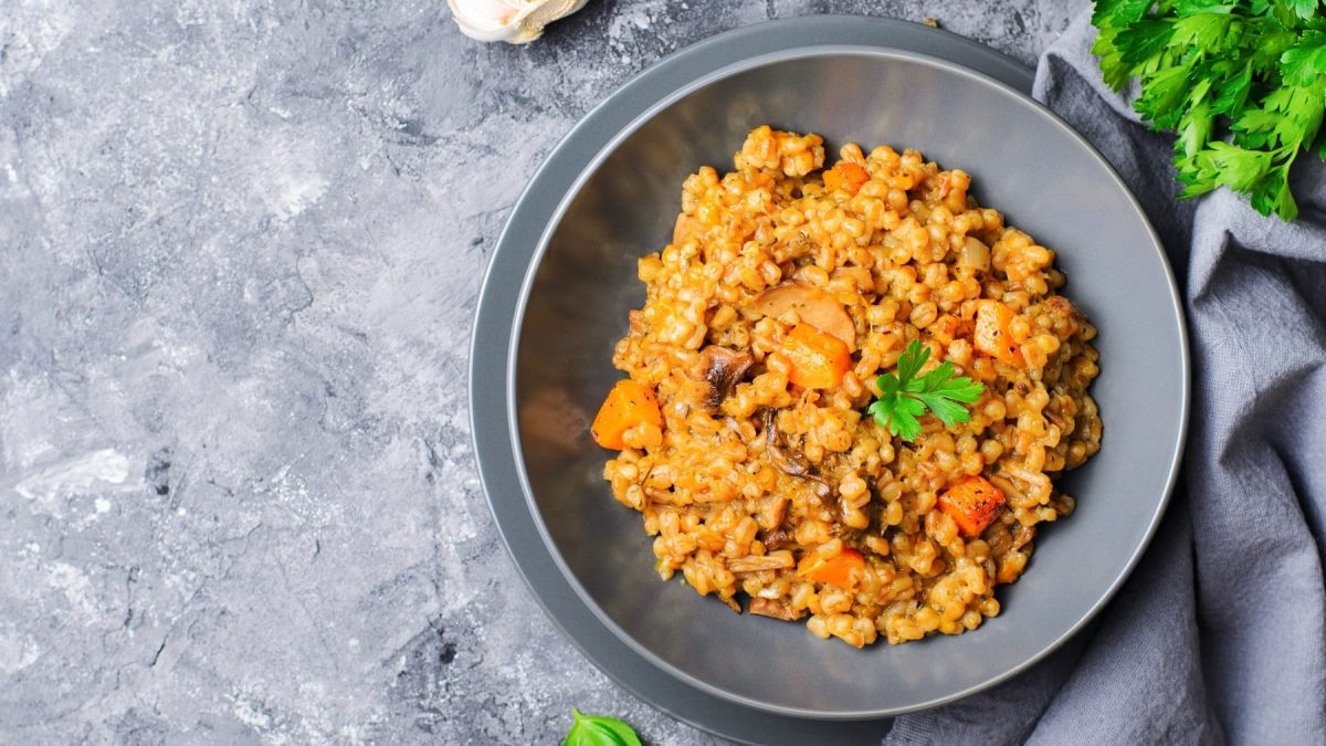 Ein grauer Teller mit dem Risotto mit Kürbis und Pilzen auf grauem Untergrund, mit grauem Tuch und Petersilie.
