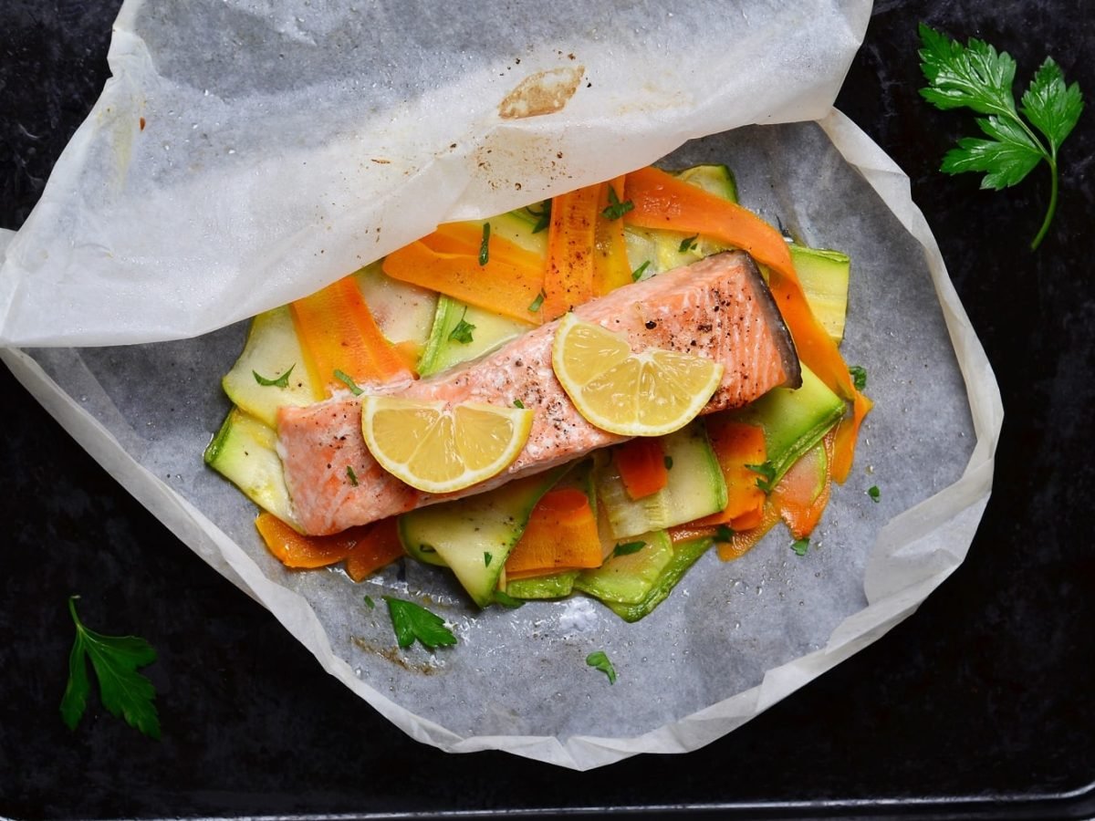 Ein schwarzes Blech mit Lachs, Gemüse und Backpapier direkt von oben auf einem dunklen Holztisch fotografiert.