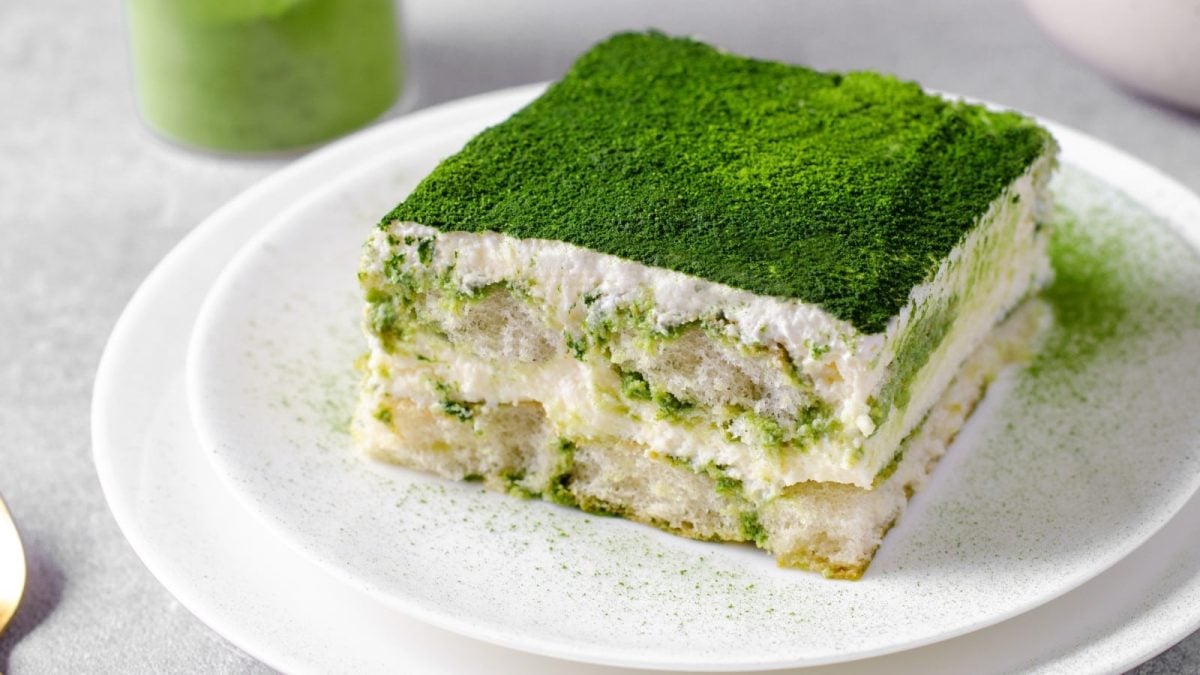 Ein Stück veganes Matcha-Tiramisu auf einem weißen Teller vor einem grauen Hintergrund. Links oben Matcha-Pulver.