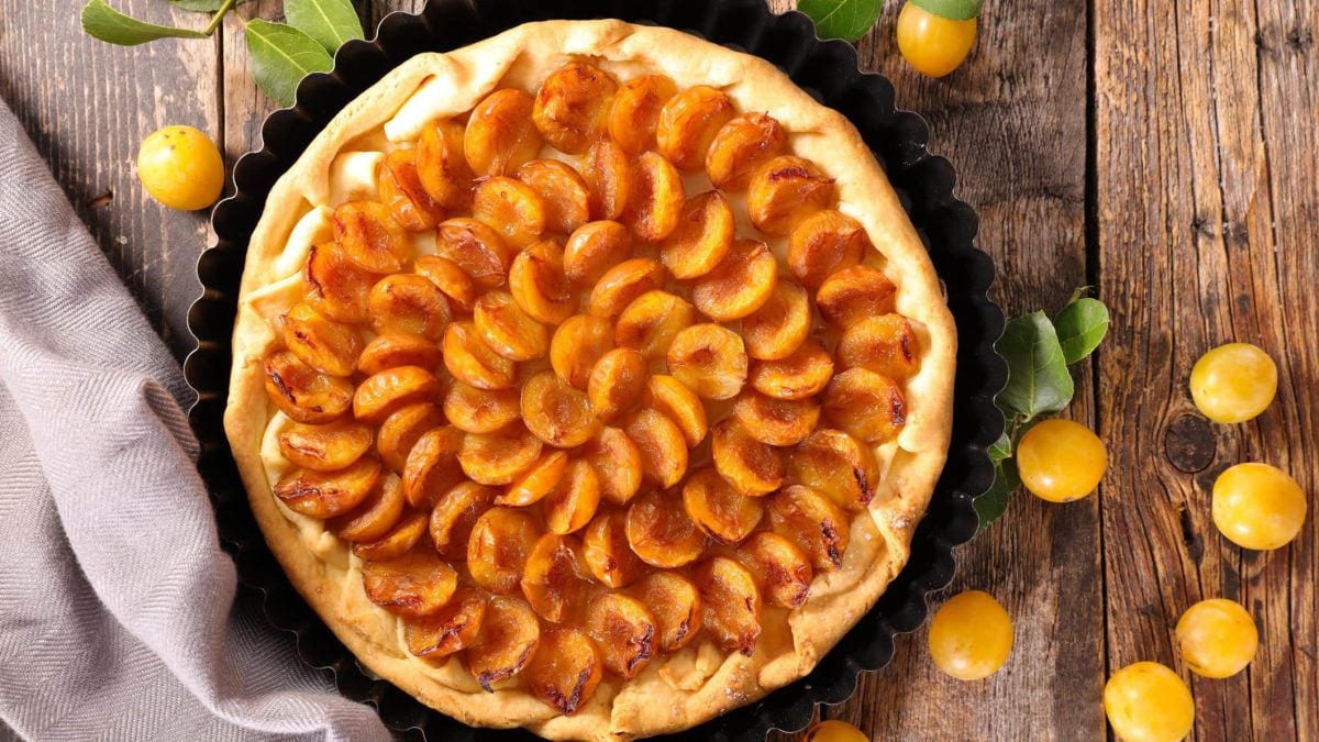 Mirabellen-Tarte mit Schmandcreme wie von Oma in schwarzer Tarteform auf Holz. Im Bild Mirabellen und ein graues Tuch. Draufsicht.