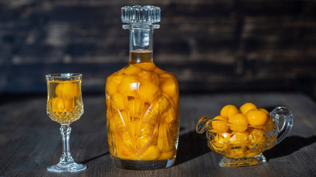 Selbst gemachter Mirabellenlikör mit ganzen Früchten in einer großen Flasche mit Glasverschluss. Daneben ganze Früchte in einer Schale und ein Glas mit Mirabellen und Mirabellenlikör. Frontalansicht.